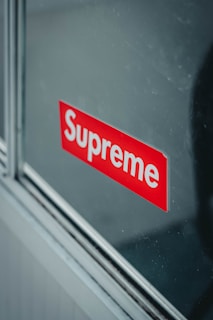 A reflective surface displays a red sticker with the brand name 'Supreme' in bold white letters. The sticker appears to be affixed onto a glass pane, with metallic or wooden trim visible along the edges.