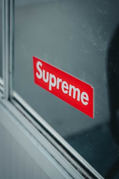 A reflective surface displays a red sticker with the brand name 'Supreme' in bold white letters. The sticker appears to be affixed onto a glass pane, with metallic or wooden trim visible along the edges.