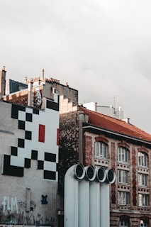 Urban scene featuring a building facade with a large pixelated black, white, and red design. Several graffiti tags are visible on the wall below. The building has multiple chimneys and there are four large industrial pipes in the foreground. A tiled roof with arched windows is visible to the right.
