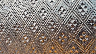 A close-up view of a glass surface with an intricate geometric pattern of diamonds and triangles. The pattern consists of repetitive diamond shapes that create a three-dimensional effect. The glass appears textured and reflects light, giving an amber tint in some areas.