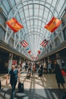 A spacious, well-lit airport corridor lined with various international flags hanging from the ceiling. People of diverse backgrounds are walking, carrying luggage, and pushing strollers. The area is bustling with travelers and the glass ceiling allows natural light to flood the space.