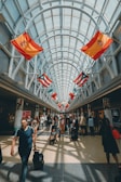 A spacious, well-lit airport corridor lined with various international flags hanging from the ceiling. People of diverse backgrounds are walking, carrying luggage, and pushing strollers. The area is bustling with travelers and the glass ceiling allows natural light to flood the space.