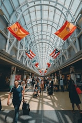 A spacious, well-lit airport corridor lined with various international flags hanging from the ceiling. People of diverse backgrounds are walking, carrying luggage, and pushing strollers. The area is bustling with travelers and the glass ceiling allows natural light to flood the space.