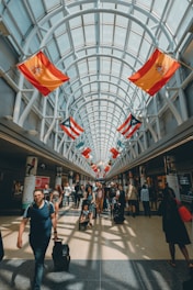 A spacious, well-lit airport corridor lined with various international flags hanging from the ceiling. People of diverse backgrounds are walking, carrying luggage, and pushing strollers. The area is bustling with travelers and the glass ceiling allows natural light to flood the space.