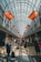 A spacious, well-lit airport corridor lined with various international flags hanging from the ceiling. People of diverse backgrounds are walking, carrying luggage, and pushing strollers. The area is bustling with travelers and the glass ceiling allows natural light to flood the space.