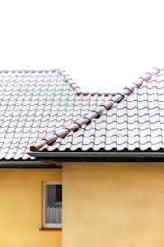 A residential building with a yellow exterior wall and a window with white curtains. The building has a tiled roof covered partially with snow, which adds a contrast between the white snow and the red-brown roofing tiles.