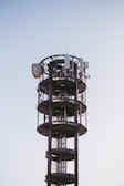 A tall, cylindrical communication tower with various antennas and satellite dishes attached to its structure, set against a clear blue sky.