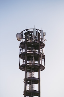 A tall, cylindrical communication tower with various antennas and satellite dishes attached to its structure, set against a clear blue sky.