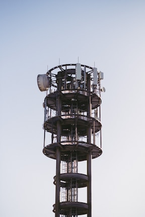 A tall, cylindrical communication tower with various antennas and satellite dishes attached to its structure, set against a clear blue sky.