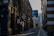 A narrow urban street surrounded by tall, brick buildings with modern and industrial architectural elements. There is a 'For Sale' sign on one of the buildings and the street is lined with street lamps. The pavement is empty, suggesting a quiet moment in the city.