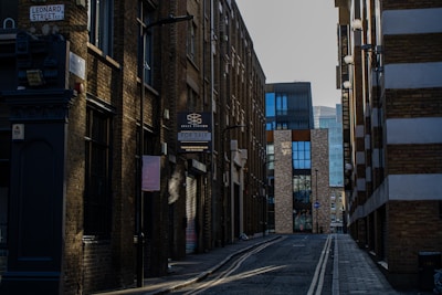 A narrow urban street surrounded by tall, brick buildings with modern and industrial architectural elements. There is a 'For Sale' sign on one of the buildings and the street is lined with street lamps. The pavement is empty, suggesting a quiet moment in the city.