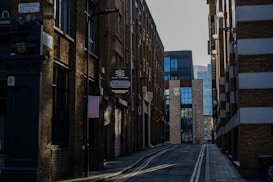 A narrow urban street surrounded by tall, brick buildings with modern and industrial architectural elements. There is a 'For Sale' sign on one of the buildings and the street is lined with street lamps. The pavement is empty, suggesting a quiet moment in the city.