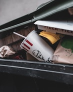 A close-up view of an open trash bin containing a fast food cup with a straw, partially crushed pizza boxes, and various paper and cardboard packaging materials.