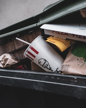 A close-up view of an open trash bin containing a fast food cup with a straw, partially crushed pizza boxes, and various paper and cardboard packaging materials.