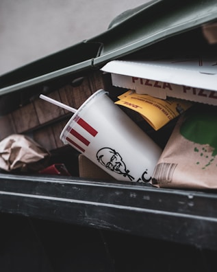 A close-up view of an open trash bin containing a fast food cup with a straw, partially crushed pizza boxes, and various paper and cardboard packaging materials.
