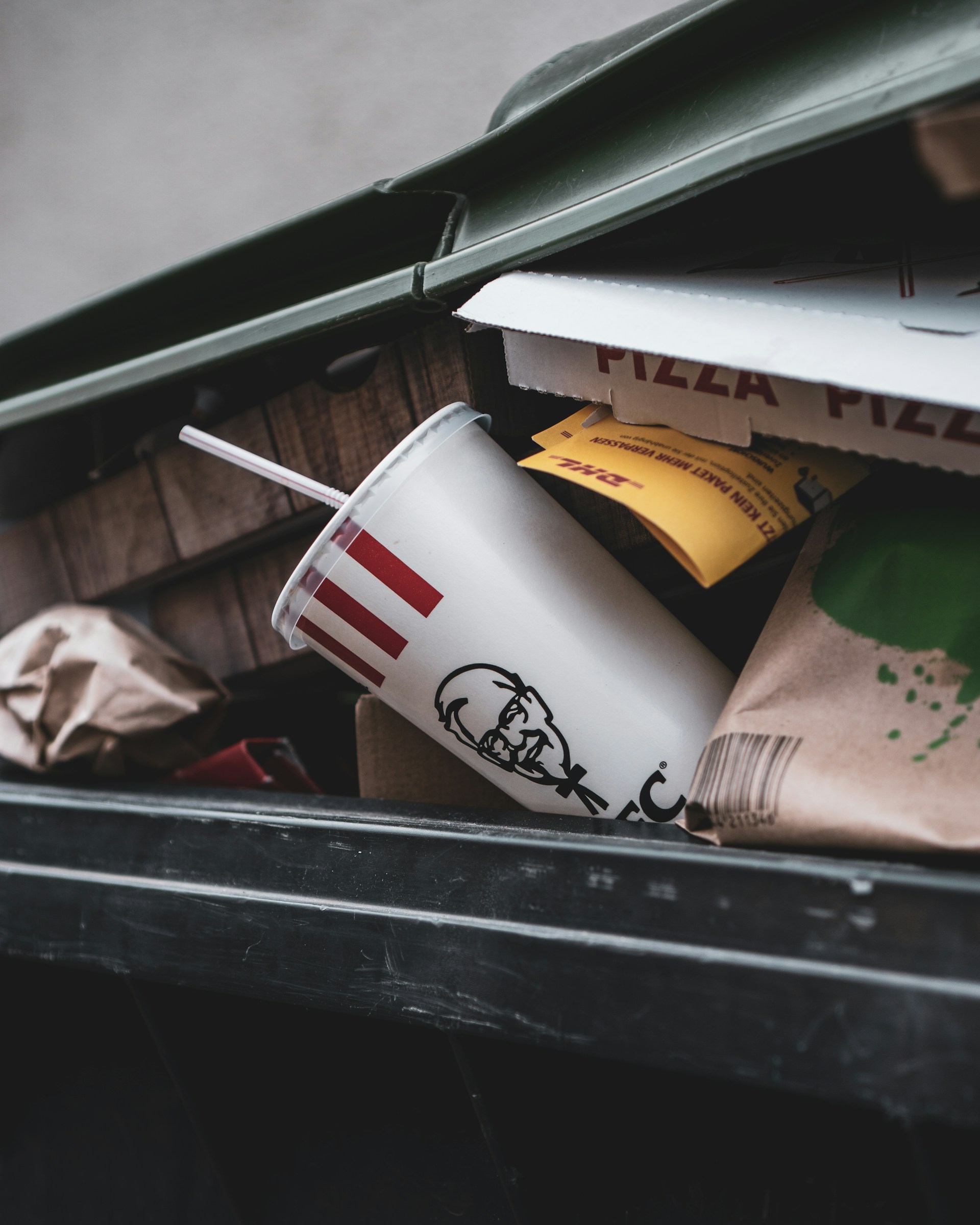 A close-up view of an open trash bin containing a fast food cup with a straw, partially crushed pizza boxes, and various paper and cardboard packaging materials.
