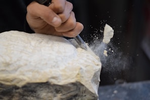 A hand is using a chisel to carve a piece of stone, with small fragments and dust flying off from the impact.