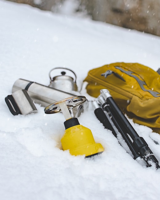 A collection of outdoor equipment is scattered on the snow, including a yellow backpack, a silver thermos, a kettle, and poles with black handles. Snow covers the ground, creating a cold and serene atmosphere.