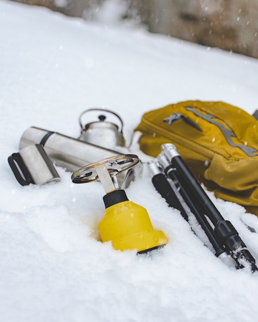 A collection of outdoor equipment is scattered on the snow, including a yellow backpack, a silver thermos, a kettle, and poles with black handles. Snow covers the ground, creating a cold and serene atmosphere.