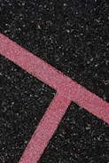 A textured black asphalt surface intersected by a bright pink line. The line is speckled with white granules, forming a distinct angular shape that stands out against the rough, dark background.