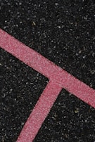 A textured black asphalt surface intersected by a bright pink line. The line is speckled with white granules, forming a distinct angular shape that stands out against the rough, dark background.