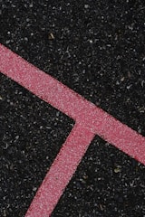 A textured black asphalt surface intersected by a bright pink line. The line is speckled with white granules, forming a distinct angular shape that stands out against the rough, dark background.