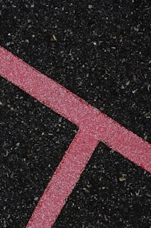 A textured black asphalt surface intersected by a bright pink line. The line is speckled with white granules, forming a distinct angular shape that stands out against the rough, dark background.