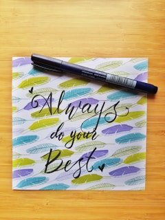 A calligraphy pen rests diagonally across a piece of paper with the motivational message 'Always do your Best' written in elegant black script. Surrounding the text are colorful feather patterns in shades of blue, green, and yellow, all set against a light wooden background.