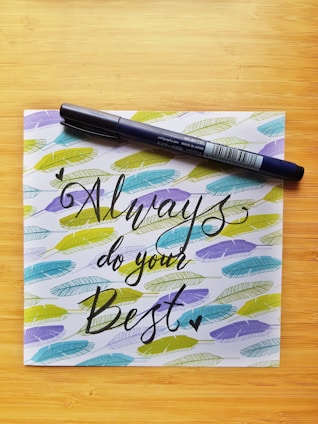 A calligraphy pen rests diagonally across a piece of paper with the motivational message 'Always do your Best' written in elegant black script. Surrounding the text are colorful feather patterns in shades of blue, green, and yellow, all set against a light wooden background.