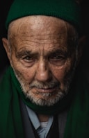 An elderly man with a deeply lined face looks intently at the viewer. He is wearing a green knit cap and a matching green scarf. His eyes convey a sense of wisdom and life experience.