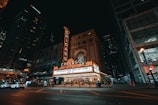 A nighttime city scene featuring a brightly lit theater marquee with the word 'Chicago' prominently displayed in neon lights. The historic architecture of the theater building blends with the modern city environment. Pedestrians and parked cars are visible in the foreground, and tall skyscrapers frame the scene.