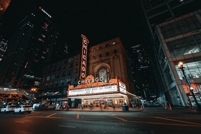 A nighttime city scene featuring a brightly lit theater marquee with the word 'Chicago' prominently displayed in neon lights. The historic architecture of the theater building blends with the modern city environment. Pedestrians and parked cars are visible in the foreground, and tall skyscrapers frame the scene.