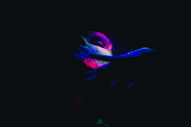 A person is illuminated by neon colors against a dark background, with vivid splashes of pink, blue, and green enhancing the facial features. The dramatic lighting casts shadows on their face and hand, creating a mysterious and artistic effect.