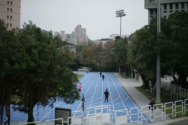 An outdoor running track with a bright blue surface curves through a park-like area. Several individuals are jogging or walking along the track. Tall buildings can be seen in the background, surrounded by trees and greenery. The scene has a cloudy sky and lacks direct sunlight, giving it a muted look.