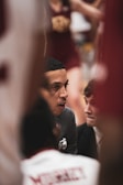 A person, likely a coach, is focused and speaking intensely to another individual. The setting appears to be a sports event, as indicated by the presence of a cheerleader in the background and someone wearing a sports jersey. The atmosphere suggests a serious discussion, possibly during a game timeout.