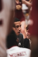 A person, likely a coach, is focused and speaking intensely to another individual. The setting appears to be a sports event, as indicated by the presence of a cheerleader in the background and someone wearing a sports jersey. The atmosphere suggests a serious discussion, possibly during a game timeout.