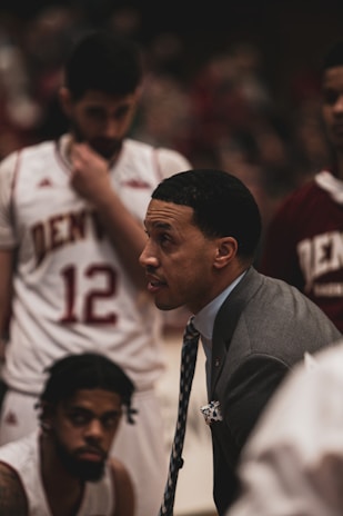 A basketball coach is in an intense discussion with his team. He is wearing a gray suit and tie, leaning forward with a focused expression. The players surrounding him are in white and maroon jerseys with 'Denver' printed on them. One player is sitting and listening attentively while another stands with a pensive look.