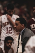 A basketball coach is in an intense discussion with his team. He is wearing a gray suit and tie, leaning forward with a focused expression. The players surrounding him are in white and maroon jerseys with 'Denver' printed on them. One player is sitting and listening attentively while another stands with a pensive look.