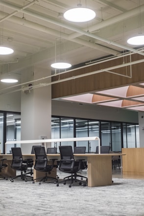 An open office space with several black swivel chairs placed around a long wooden table with light grey carpet on the floor. The ceiling design features exposed pipes and round white ceiling lights which create a modern, industrial look. Large glass walls line one side of the room.