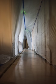 A long, empty hallway partially covered with white plastic sheets draped from the ceiling, giving the impression of a construction or renovation site. The floor is reflective, suggesting a polished surface.