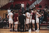 A group of basketball players in white uniforms huddle together on a wooden court, with a coach in a dark suit facilitating the meeting. Two players have their backs to the camera, showing numbers 12 and 21. Spectators in casual clothing fill the stands in the background, some seated and others standing.