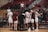 A group of basketball players in white uniforms huddle together on a wooden court, with a coach in a dark suit facilitating the meeting. Two players have their backs to the camera, showing numbers 12 and 21. Spectators in casual clothing fill the stands in the background, some seated and others standing.