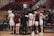 A group of basketball players in white uniforms huddle together on a wooden court, with a coach in a dark suit facilitating the meeting. Two players have their backs to the camera, showing numbers 12 and 21. Spectators in casual clothing fill the stands in the background, some seated and others standing.