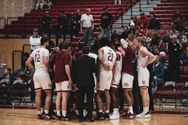 A group of basketball players in white uniforms huddle together on a wooden court, with a coach in a dark suit facilitating the meeting. Two players have their backs to the camera, showing numbers 12 and 21. Spectators in casual clothing fill the stands in the background, some seated and others standing.