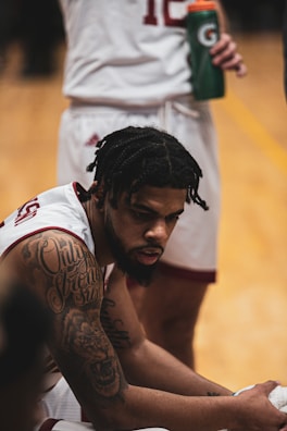 A basketball player with elaborate tattoos on his arm is seated on the bench, appearing focused or deep in thought. He wears a white jersey with maroon accents. In the background, another player stands holding a sports drink bottle.