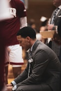 A man in a suit is kneeling, appearing to be focused or in thought. Around him is a standing person wearing a maroon sports shirt with partially visible text. The setting seems to be related to sports, possibly a basketball game or practice, with a clipboard held by another person in the background.
