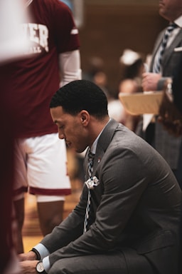 A man in a suit is kneeling, appearing to be focused or in thought. Around him is a standing person wearing a maroon sports shirt with partially visible text. The setting seems to be related to sports, possibly a basketball game or practice, with a clipboard held by another person in the background.