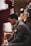 A man in a suit is kneeling, appearing to be focused or in thought. Around him is a standing person wearing a maroon sports shirt with partially visible text. The setting seems to be related to sports, possibly a basketball game or practice, with a clipboard held by another person in the background.