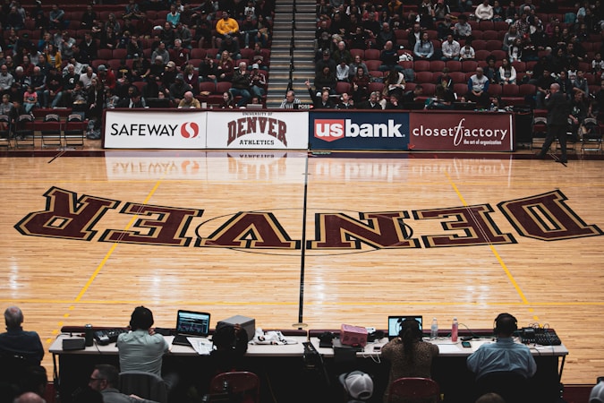 A basketball court with the word 'DENVER' prominently displayed on the polished wood floor. People are seated in the background on red chairs, observing or waiting for an event. Advertisements for Safeway, US Bank, and Closet Factory are visible above the court. Four individuals are seated at a table in the foreground, working with electronic devices.