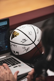 A signed basketball rests against a laptop on a table. A person's hand is visible, possibly using the laptop, and a microphone can be seen close by, suggesting an interview or commentary setting.
